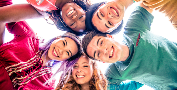 A group of teenagers huddled together in a circle. They are smiling wearing colourful clothing