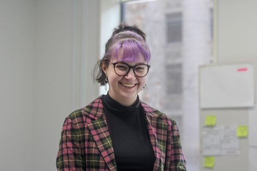 A person with purple hair, wearing glasses and a coloured checkered blazer smiling at the camera, with an office wall behind them.