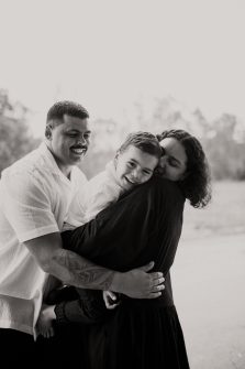 Black and white family portrait of Tanika, Adrian and their young son Slade in a field. They're warmly hugging and smiling.