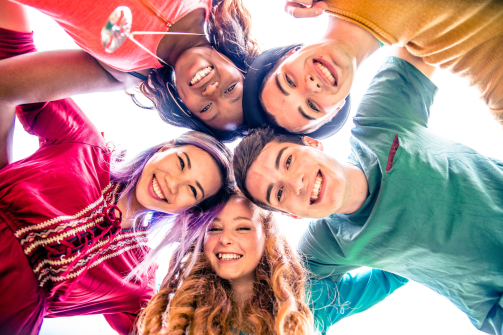 A group of teenagers standing together in a huddle. They are smiling down at the camera wearing colourful clothing. 