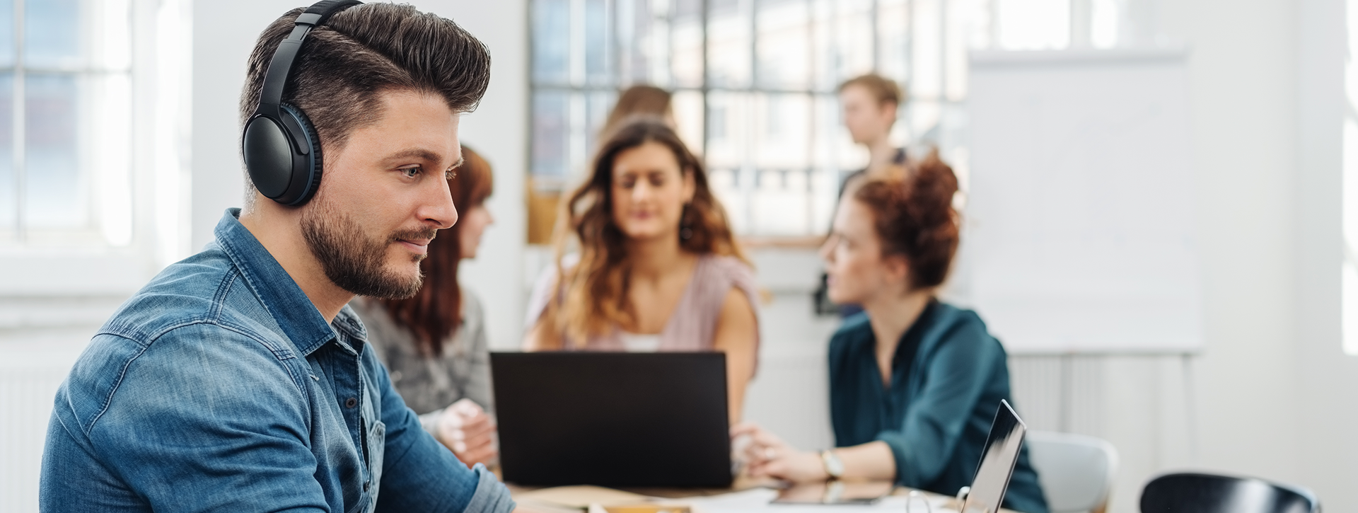 Man at workplace on computer wearing headphones. He is in a blue shirt and has short hair and a beard. Blurred in the background are other people in a working environment.