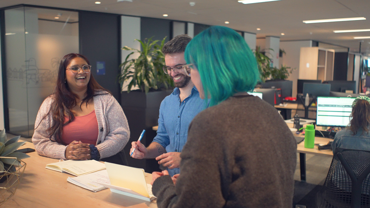 Three people sit at a desk. There are books in front of them and a succulent. There is a smiling woman in a salmon-coloured tank top and cream cardigan, a smiling man in a blue shirt who is looking down at a notebook, and a woman with teal hair who has her back to the camera. They all appear to be collaborating on a project.