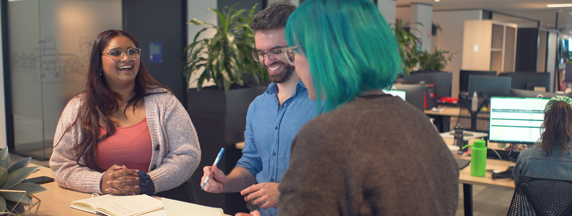 Three people sit at a desk. There are books in front of them and a succulent. There is a smiling woman in a salmon-coloured tank top and cream cardigan, a smiling man in a blue shirt who is looking down at a notebook, and a woman with teal hair who has her back to the camera. They all appear to be collaborating on a project.