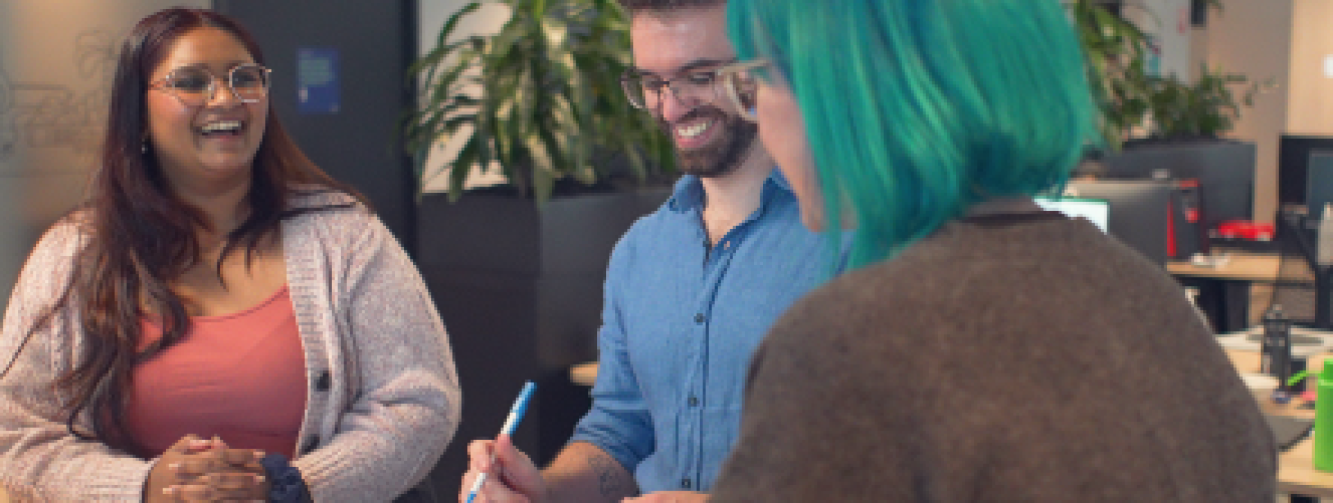 Three people sit at a desk. There are books in front of them and a succulent. There is a smiling woman in a salmon-coloured tank top and cream cardigan, a smiling man in a blue shirt who is looking down at a notebook, and a woman with teal hair who has her back to the camera. They all appear to be collaborating on a project.